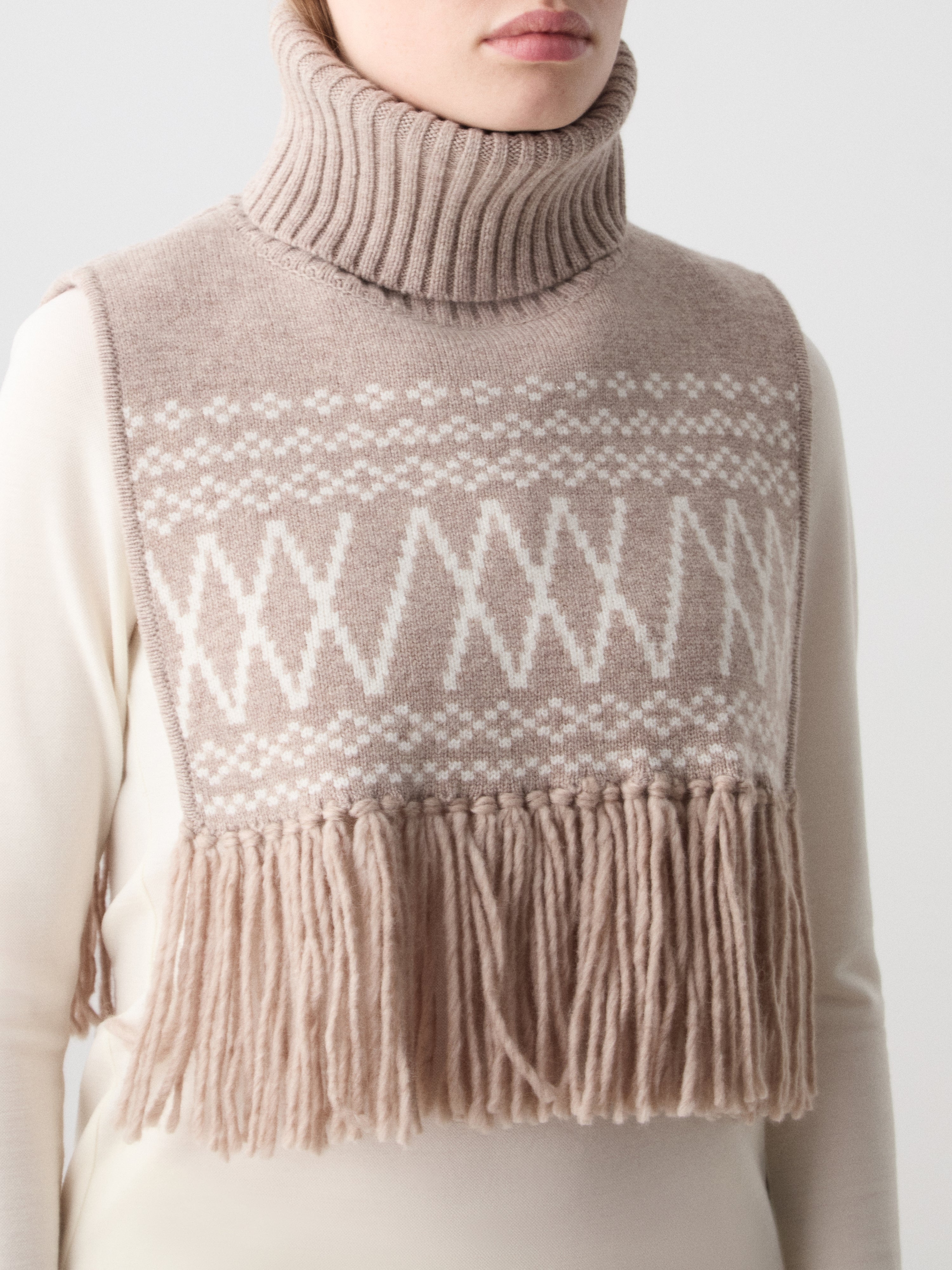 We Norwegians | FW25 | Setesdal Bib_Sand | Model is wearing the knitted bib with a turtle neck and fine fringes at the hem over a white sweater, against an off-white background