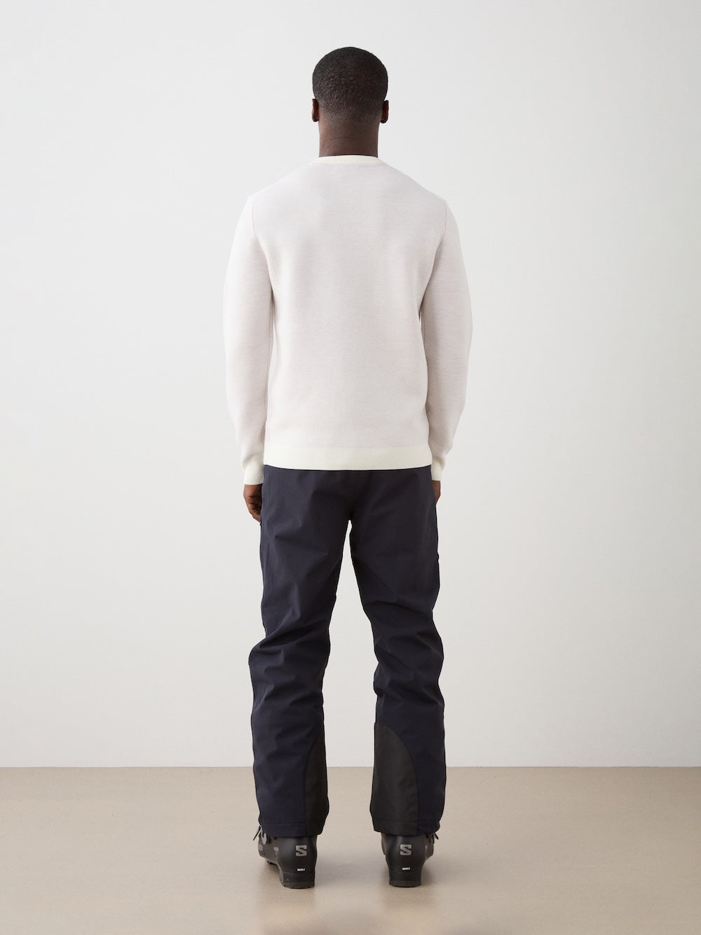 We Norwegians | FW25 | Mens Afterski Crewneck_Cream | Model wearing a knitted sweater with a design and logo at the front with black ski pants | Shot against an off-white background | Back view