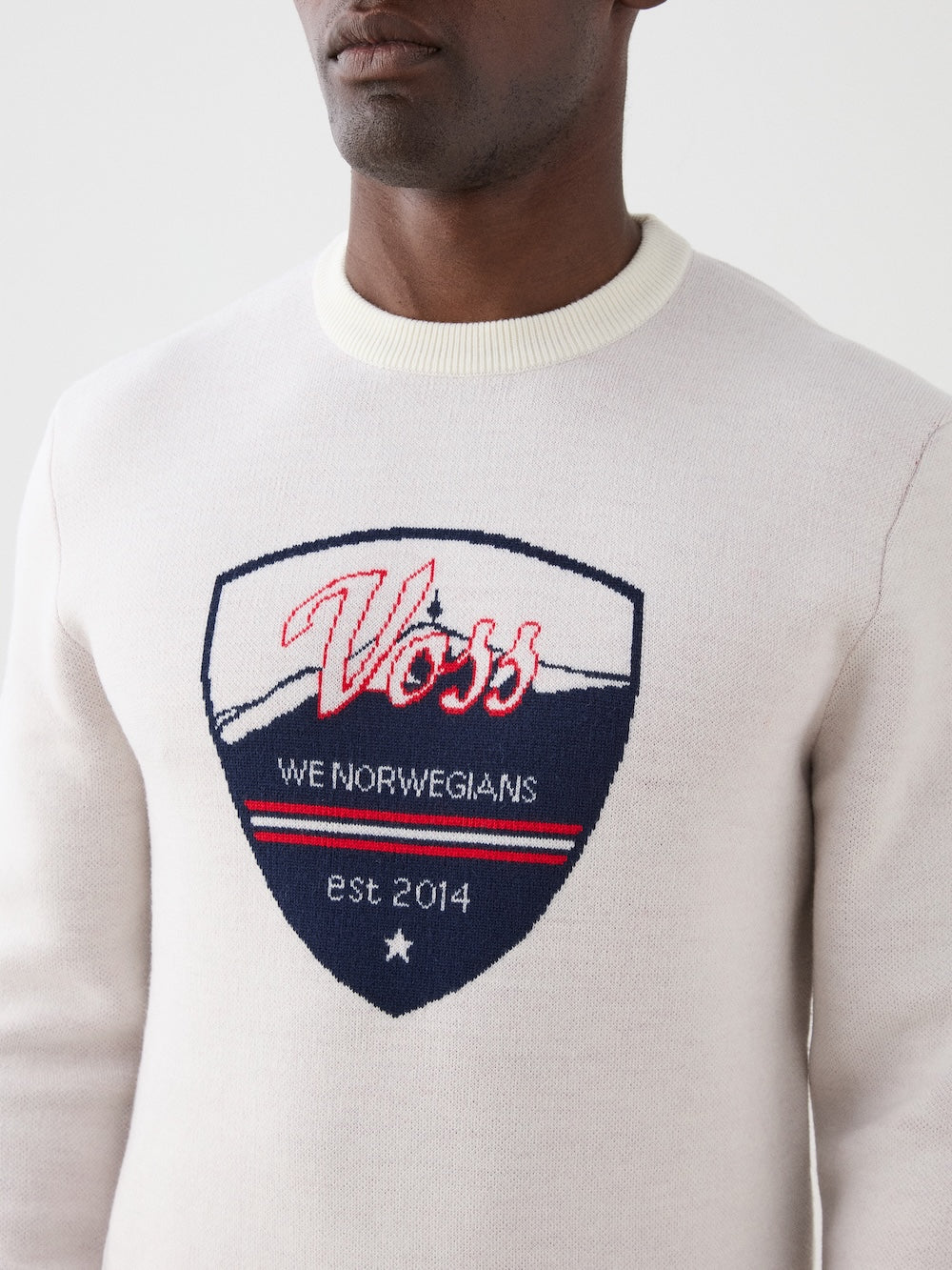 We Norwegians | FW25 | Mens Afterski Crewneck_Cream | Model wearing a knitted sweater with a design and logo at the front with black ski pants | Shot against an off-white background | Close-up view